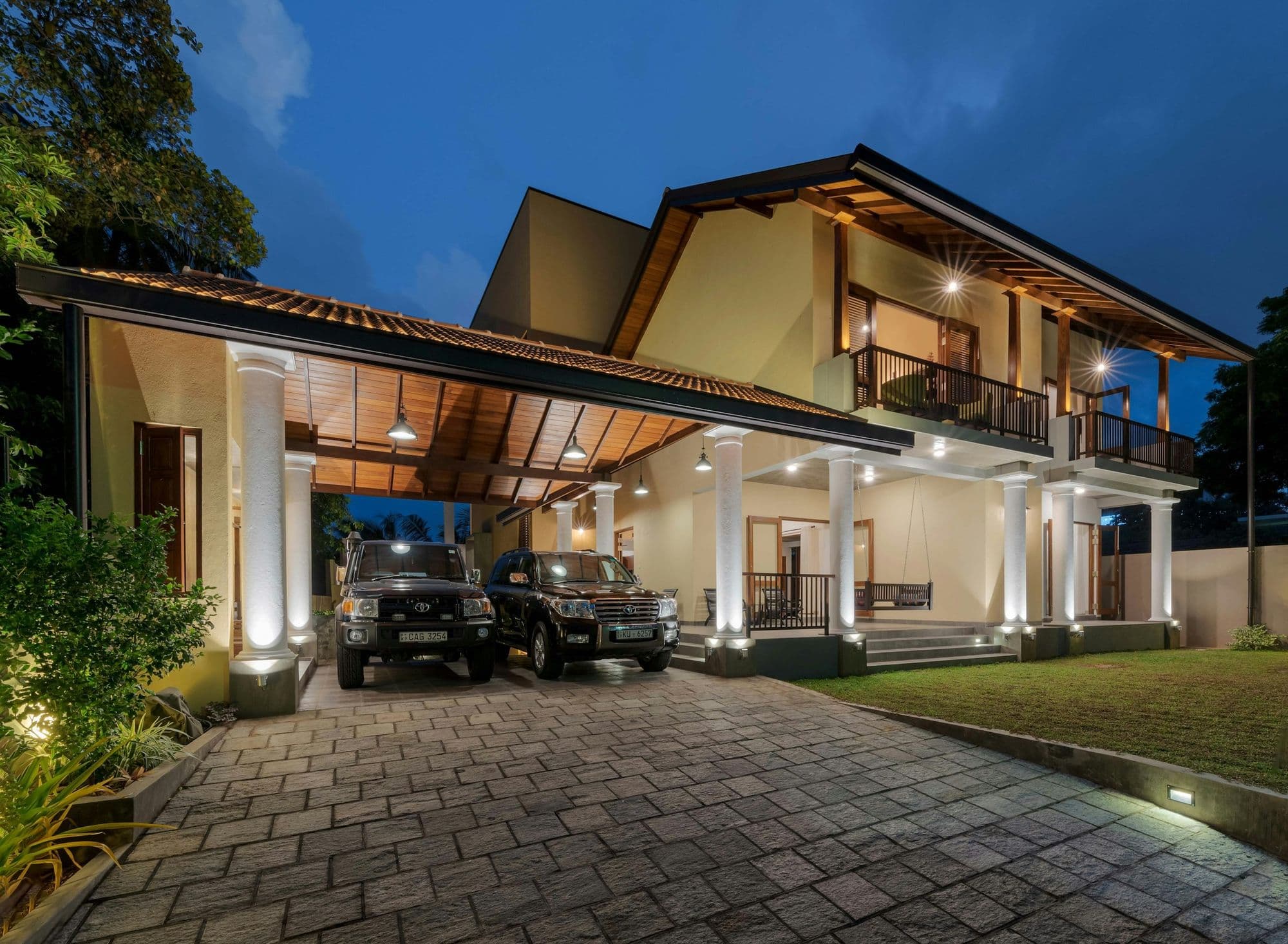A modern two-storey family home at dusk, lit from within, with two SUVs parked under the carport and a paved driveway.