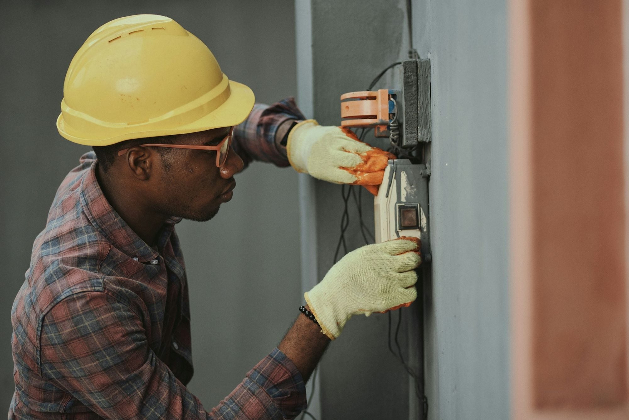 An electrician in a yellow hard hat and gloves working on a wall-mounted electricity meter.