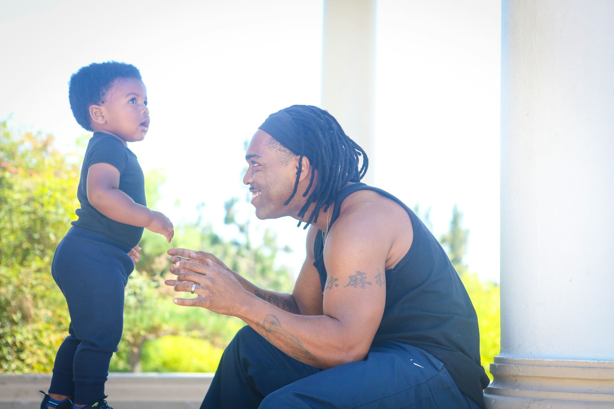 A father crouching on his porch, smiling as his toddler walks towards him.